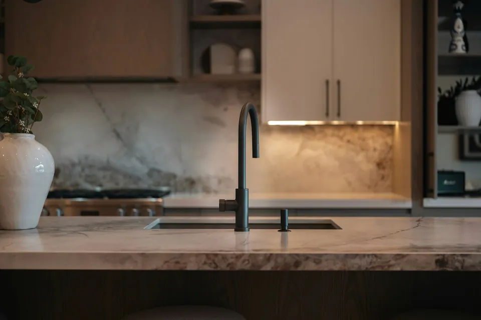Kitchen island with a dark gooseneck tap, veined stone-look worktop and pale cabinets with warm under-lighting.