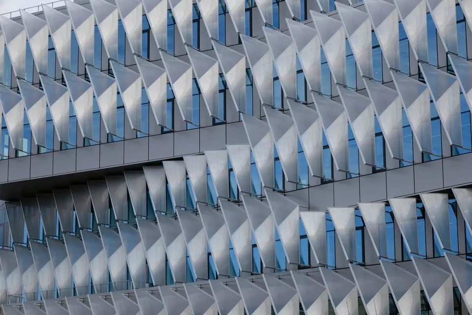 Building facade covered in rows of angled triangular white panels over blue glass windows.