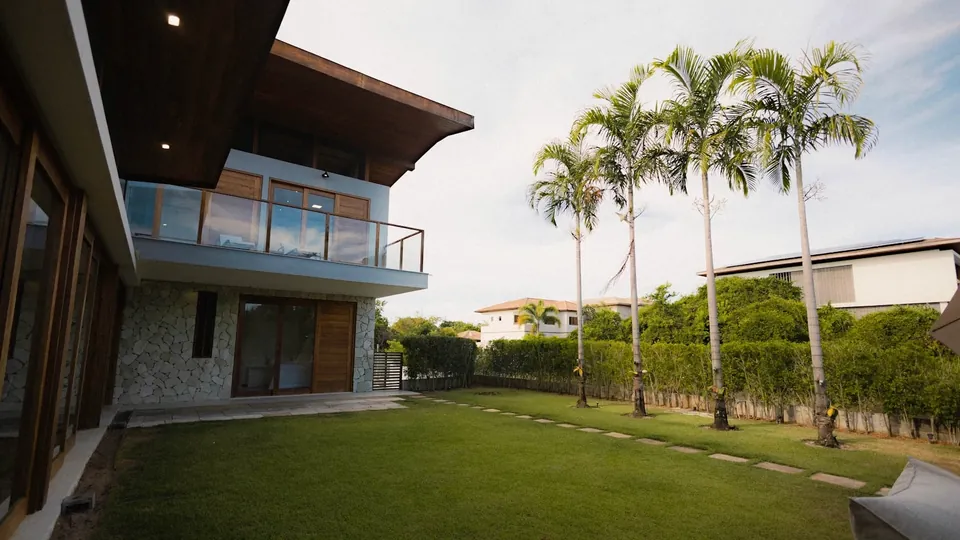 Two-storey house with a glass balcony and stone wall overlooking a lawn lined with tall palm trees.