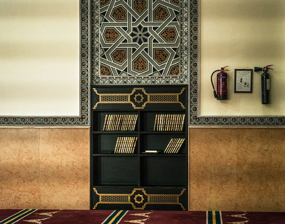 Wall with an ornate geometric star pattern panel above a dark bookcase, beside fire extinguishers on a red carpet.