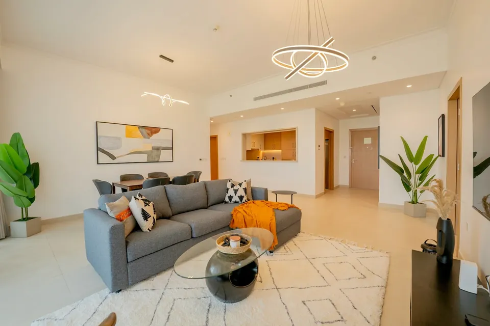 Bright living room with a grey sofa, orange throw, round glass coffee table and a dining area beyond.