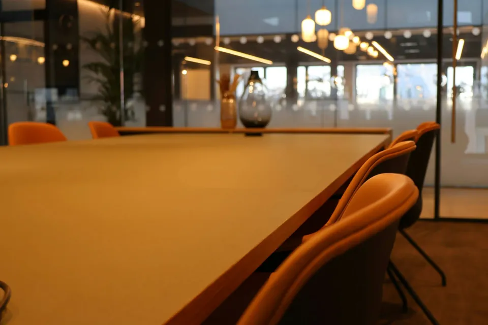 Meeting table with orange chairs in a glass-walled office lit by hanging bulb lights.
