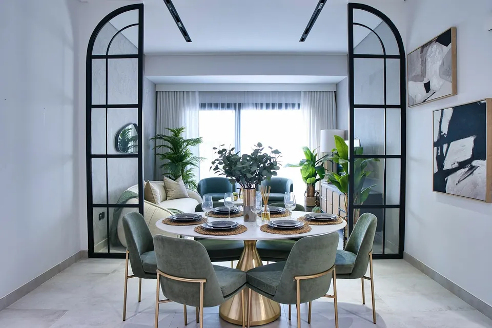 Dining room seen through black-framed arched glass doors, with green chairs around a round gold-based table.