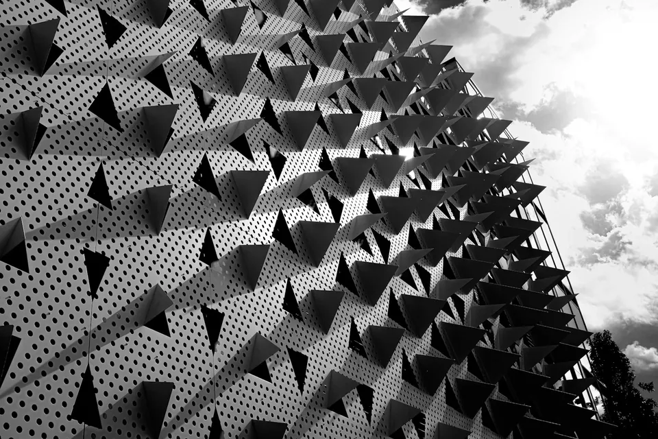 Black and white facade of perforated panels with rows of projecting triangular fins against a cloudy sky.