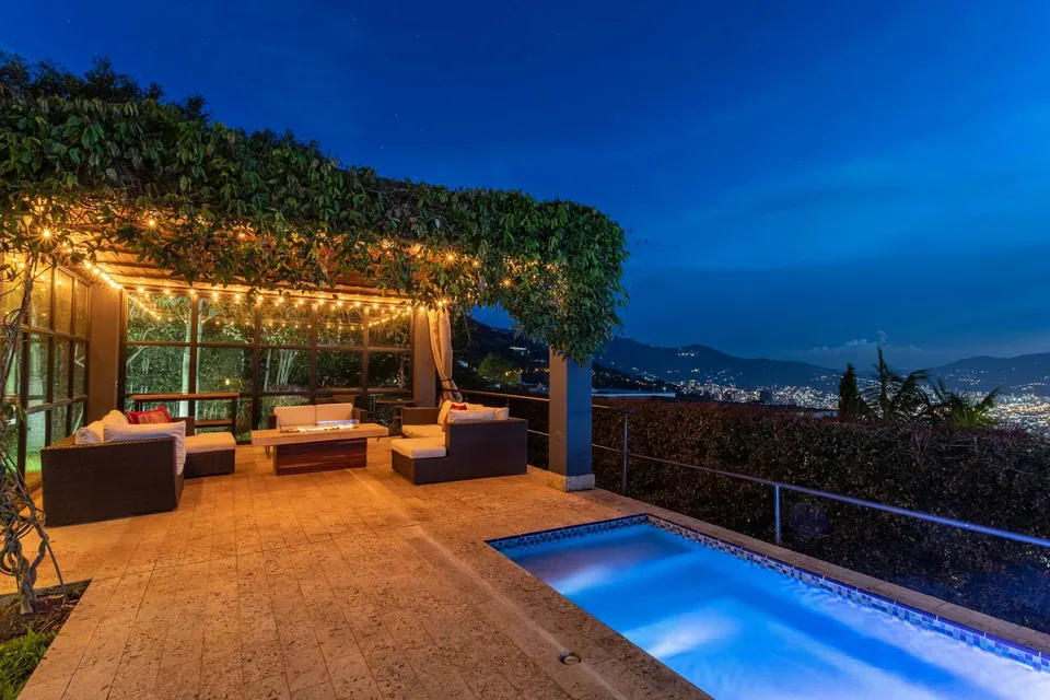 Terrace at dusk with a vine-covered pergola strung with lights, outdoor sofas and a lit pool overlooking hills.