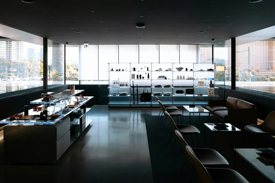 Dark retail interior with glass display counters, lit shelving along a glazed wall and chairs by low tables.