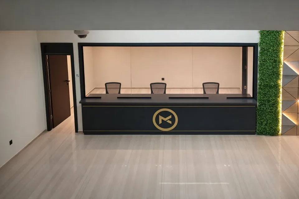 MK Pay, Al Rigga: service counter with three chairs behind glass, a gold circular logo and a green plant wall.