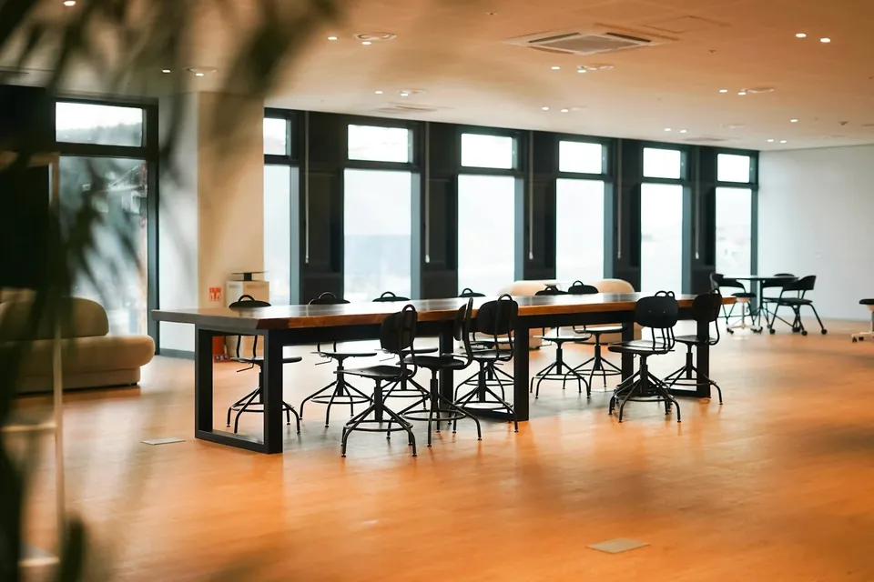 Bright open office with a long wooden table, black stools and a row of tall windows, seen past blurred leaves.