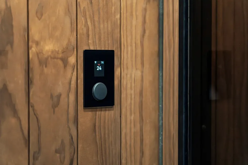 Black smart thermostat panel with a small screen and round dial mounted on a wood-panelled wall.