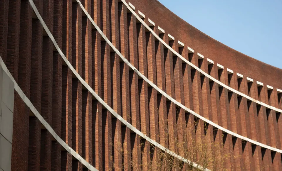 Curved red brick building facade with deep vertical fins and white horizontal bands against a blue sky.