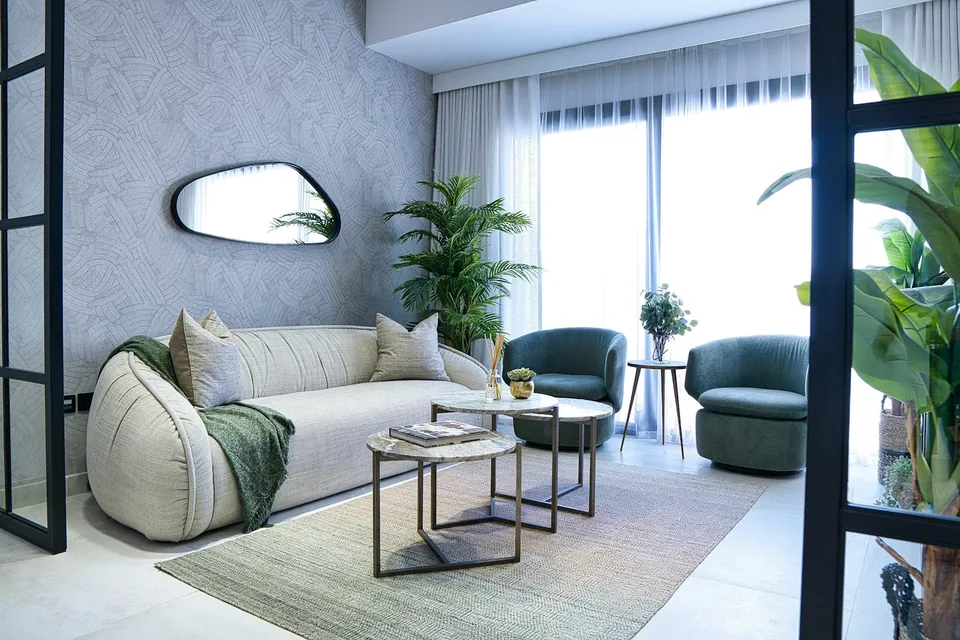 Living room with a curved cream sofa, green armchairs, nested side tables and a pebble-shaped mirror.