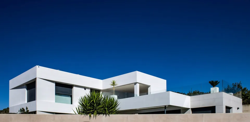 White flat-roofed modern house with stacked boxy volumes and spiky plants under a deep blue sky.