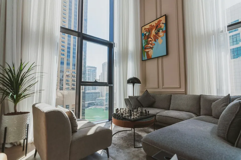 Grey sectional sofa and armchair by tall windows with high-rise views, a chess set and a colourful portrait.