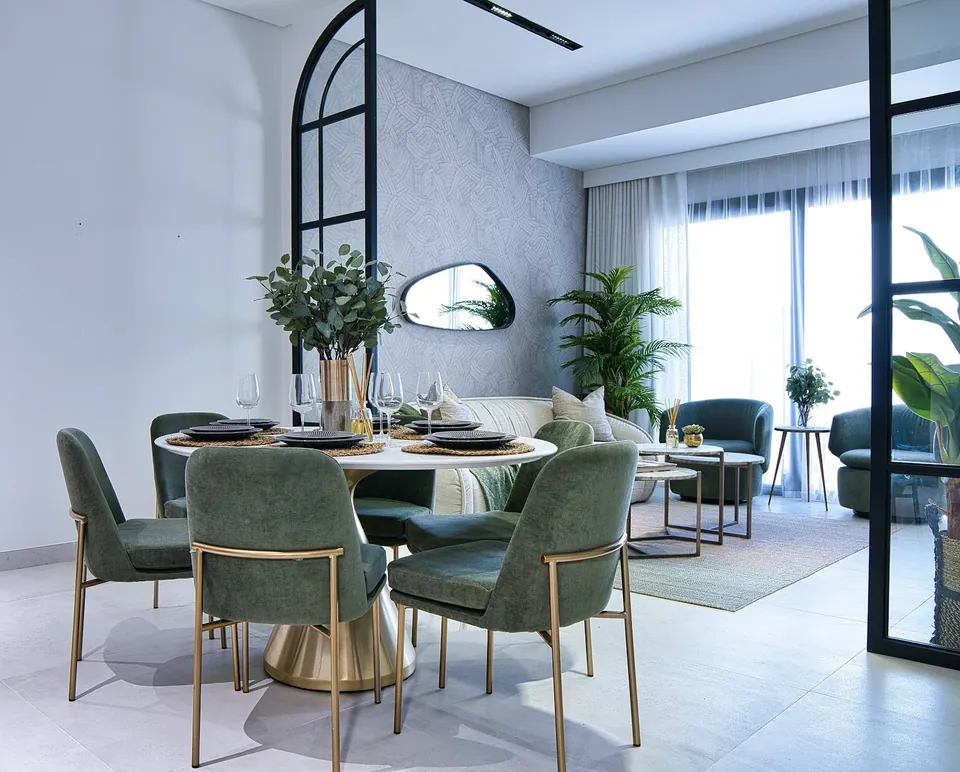 Dining area with green chairs around a round gold-based table, beside a lounge with a pebble-shaped mirror.