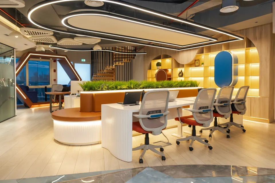 Open office with white desks, grey mesh chairs, a grass planter, lit wooden shelving and curved ceiling light bands.
